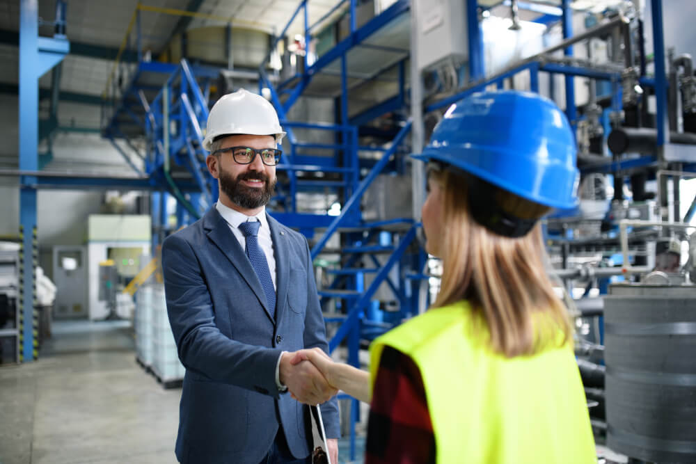 Ein Verkäufer in Anzug und Helm schüttelt einem Ingenieur in einer Fabrik die Hand, was für ein erfolgreiches Geschäft im Maschinenbau steht